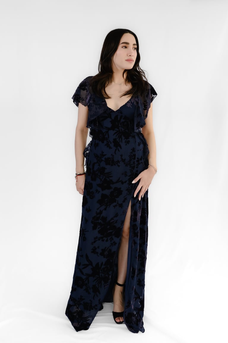 Woman wearing a dark navy velvet floral dress with short sleeves and a high slit, standing against a white background