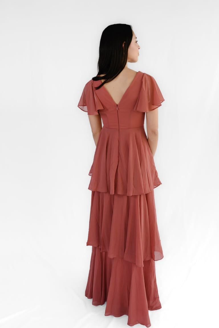 Woman wearing a rust-colored tiered chiffon dress with flutter sleeves, shown from back profile against white background