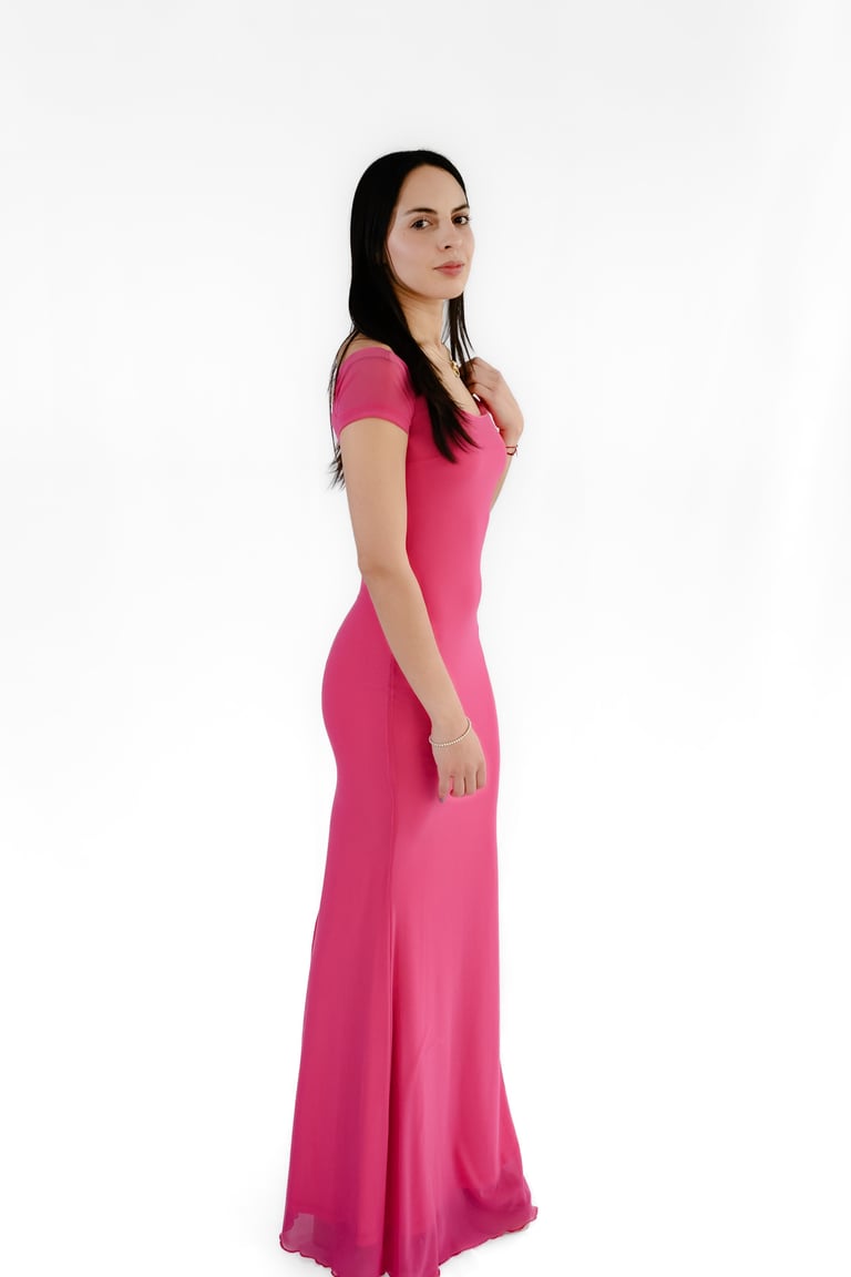 Woman with long dark hair wearing a pink floor-length gown with short sleeves, standing against a white background
