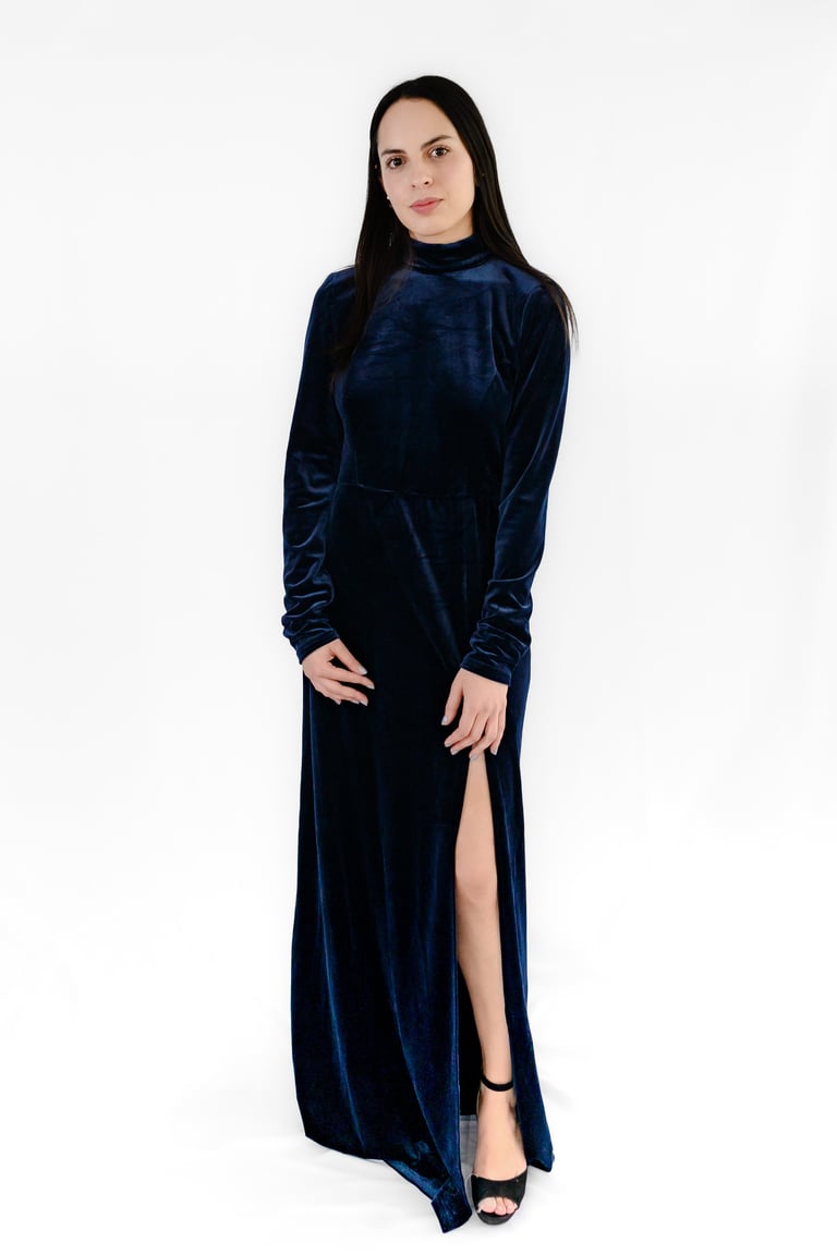 Woman wearing a dark blue velvet maxi dress with long sleeves and a high slit, standing against a white background