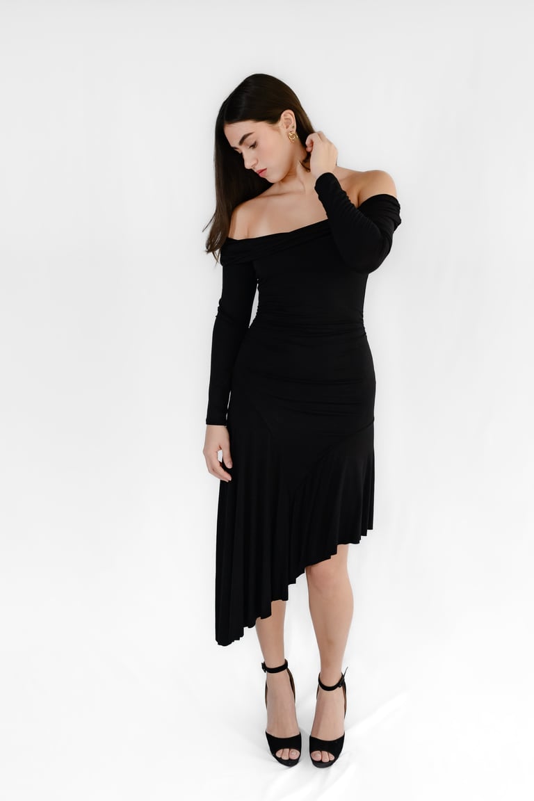 Woman in black off-shoulder dress with asymmetrical hem and black platform heels posing against white background