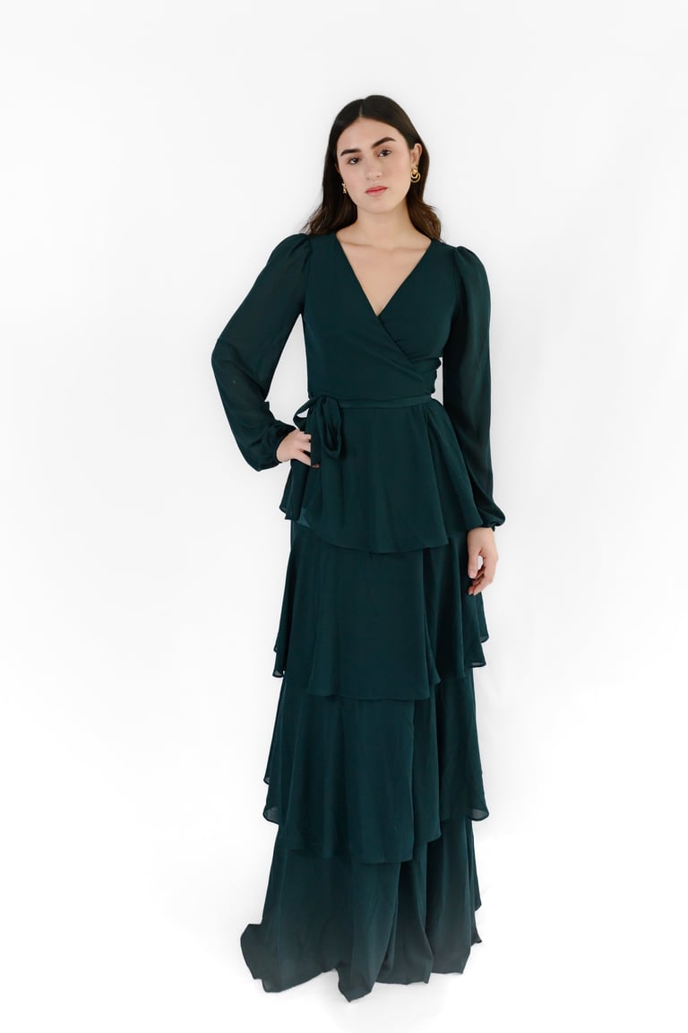 Woman wearing a dark green wrap dress with long sleeves and tiered ruffled skirt standing against a white background