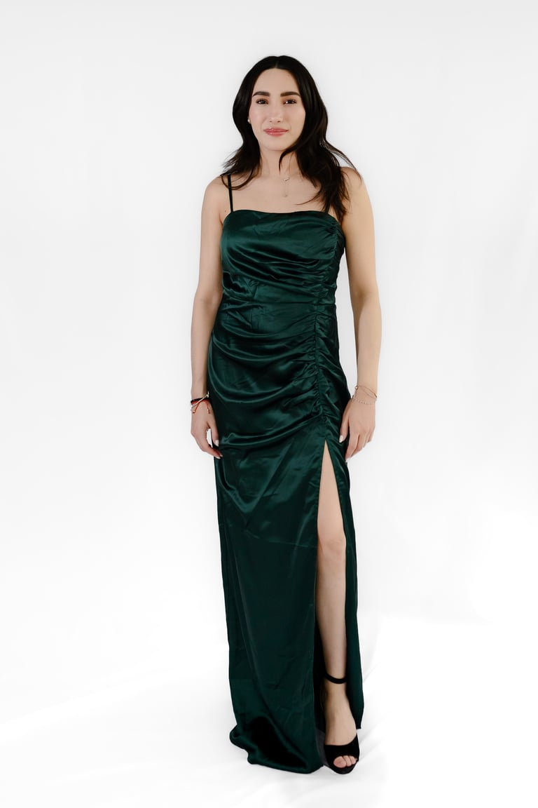 Woman wearing a dark green satin floor-length gown with ruched detailing and a high slit, standing against a white background